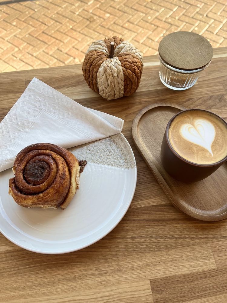 A cinnamon roll on a white plate beside a latte with rosetta art on an outdoor table