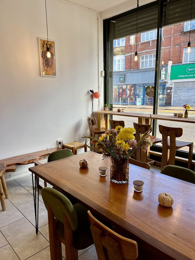 The long communal table with green chairs beside the full-height front window, dressed with yellow flowers
